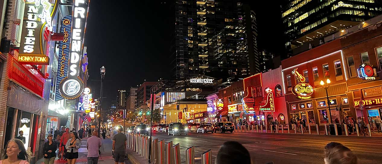 Nighttime street scene in downtown Nashville, TN with brightly lit signs of various bars and restaurants. People walk along the sidewalks, and cars drive down the street. Tall buildings are seen in the background.