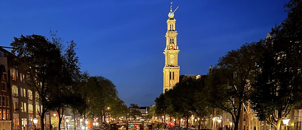 A lit-up church tower along an Amsterdam canal at dusk, surrounded by trees and buildings, with a bridge and streetlights in the foreground.