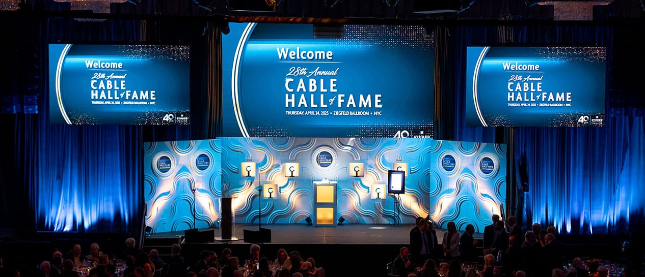 A stage with large screens displays “Cable Hall of Fame” at a formal event, with people seated at round tables in the foreground.