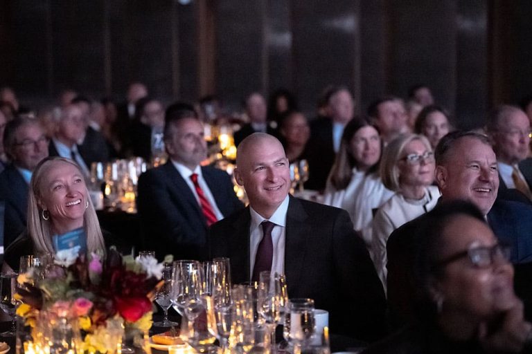 A group of people in formal attire sit at round tables with floral centerpieces and glassware, attentively watching an event or presentation.