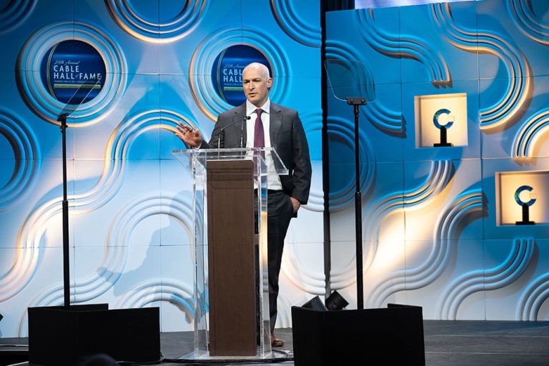 A man in a suit speaks at a clear podium on a stage with blue and white decorative panels and "Cable Hall of Fame" signs in the background.