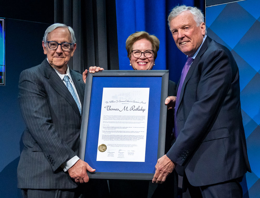 Three people in formal attire stand together, holding a large framed certificate featuring a gold seal and text, smiling for the camera—celebrating mentorship and ethical standards in the cable sector.