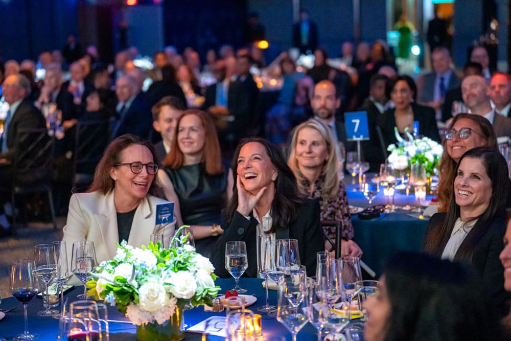 A group of well-dressed people, including 2026 honoree Kristin Dolan, CEO of AMC Global Media, sit at round tables, smiling and laughing during an indoor Cable Hall of Fame event in NYC, surrounded by floral centerpieces and wine glasses.