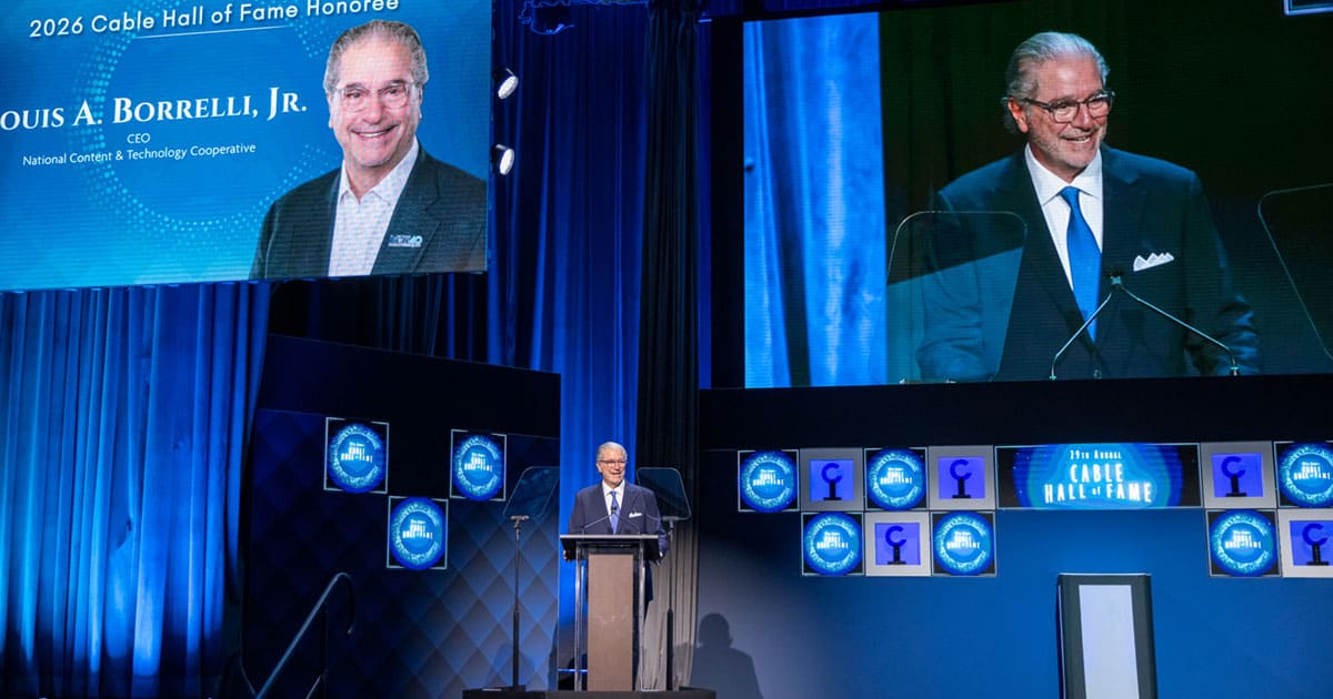 A man speaks at a podium on stage in NYC, beside screens displaying his photo and the text "2026 Cable Hall of Fame Honoree Louis A. Borrelli, Jr.", celebrating one of the esteemed 2026 honorees.