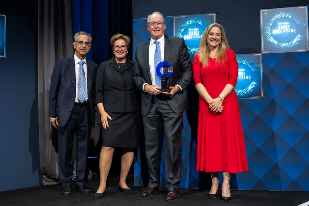 Four people in business attire stand on a stage; David M. Fellows Former CTO, Comcast Former Chair of the Cable TV Pioneers, in the center holds his 2026 Cable Hall of Fame honoree Award. Blue and white signage reads "Cable Hall of Fame NYC," celebrating the 2026 honorees in the background.